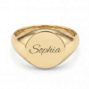 May include: A gold signet ring featuring a smooth, circular face with the name Sophia engraved in a cursive script font. The ring has a polished finish and a solid band that tapers toward the back, showcasing a classic, clean design.