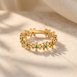 May include: A gold-colored ring featuring a daisy flower design. Each flower has a turquoise center. The ring is set against a neutral background, highlighting the jewelry's intricate details. The ring is a delicate piece of fashion jewelry.