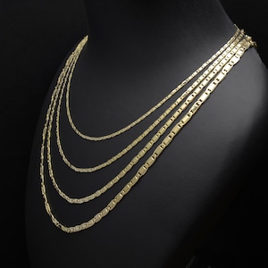 May include: A gold-colored necklace set displayed on a black mannequin. The set includes multiple strands of varying lengths, each featuring a flat, rectangular link design. The necklaces are arranged to cascade down, showcasing their layered effect.