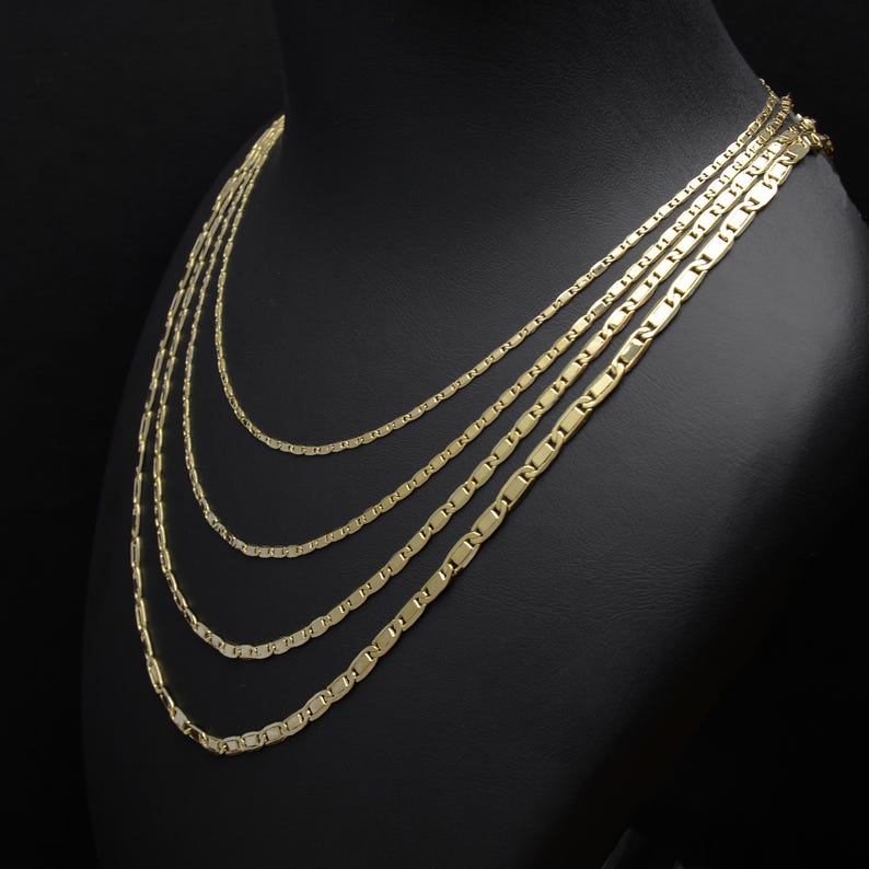 May include: A gold-colored necklace set displayed on a black mannequin. The set includes multiple strands of varying lengths, each featuring a flat, rectangular link design. The necklaces are arranged to cascade down, showcasing their layered effect.