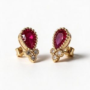 May include: A pair of gold stud earrings featuring a central pear-shaped red gemstone surrounded by a beaded metal border, accented with three small clear round stones at the base. The earrings include standard post backs for secure attachment.