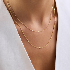 May include: A double-layered gold chain necklace featuring delicate, thin links interspersed with small, flat, rectangular metallic accents. The two chains hang at slightly different lengths, creating a layered appearance against a white garment.