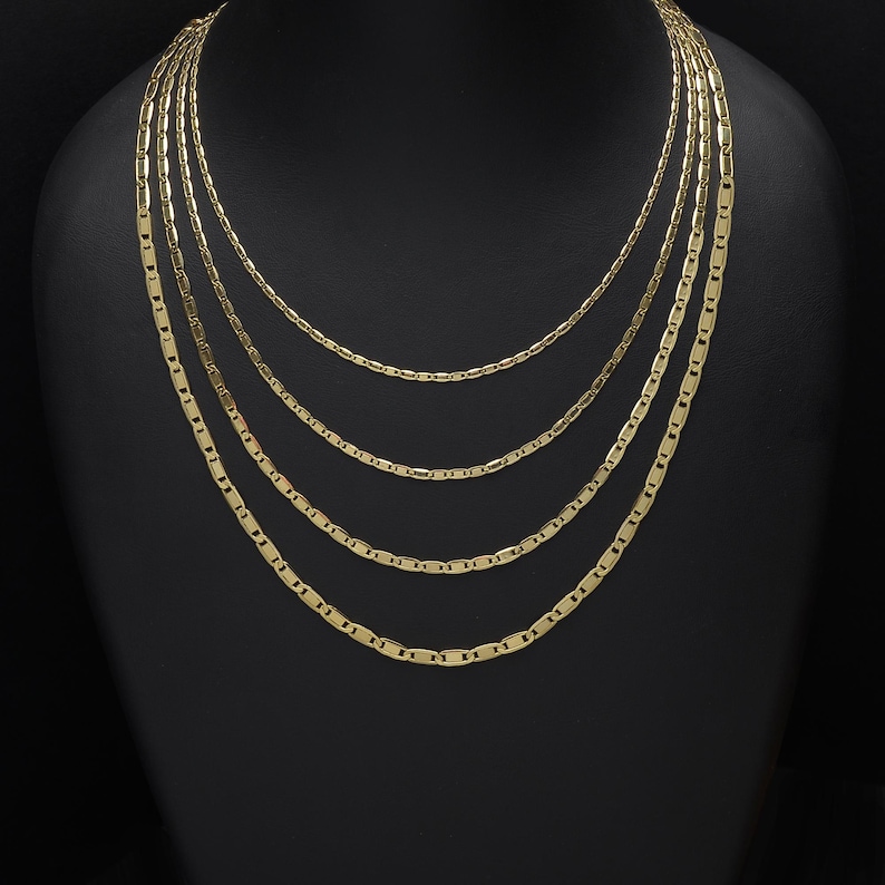May include: A display of layered gold necklaces. The necklaces have a flat, rectangular link design. The chains are displayed on a black mannequin, showcasing the different lengths and styles of the jewelry.