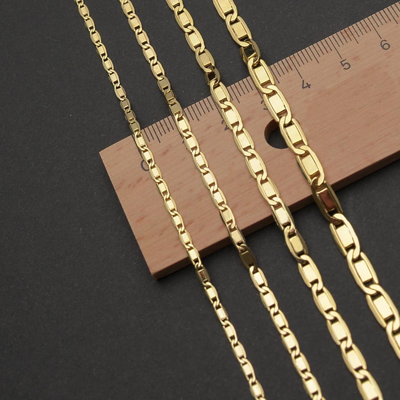 May include: Close-up of several gold-colored chain necklaces. The chains have a flat, rectangular link design. A wooden ruler with inch markings is partially visible, providing scale. The background is a dark, solid color.
