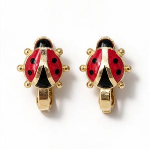 May include: A pair of gold-tone clip-on earrings shaped like ladybugs. Each earring features a bright red enamel back with black spots, a black enamel head, and gold-tone metal accents along the edges and sides, including small rounded protrusions.