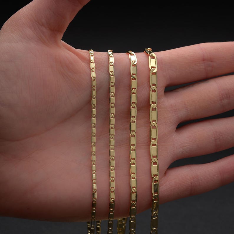 May include: Four gold-colored chain necklaces of varying thicknesses are displayed. The chains have a rectangular link design. The chains are held in a hand against a dark background.