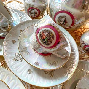 May include: A porcelain tea set featuring an iridescent finish, gold filigree accents, and central medallions with colorful pastoral scenes framed in magenta. The set includes a teapot, a lidded sugar bowl, multiple teacups, and matching saucers.
