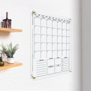 Personalized acrylic wall calendar with gold hardware, featuring sections for notes, menu, and individual family member schedules, set against a warm home setting.