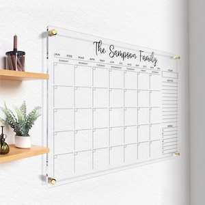 Clear acrylic wall calendar with gold hardware, featuring a full monthly layout, sections for year box, notes, and menu, displayed on a bright wall next to a decorative shelf.