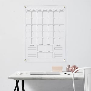 Featuring a monthly layout, notes, a menu section, and personalized spaces for four name boxes. Clear acrylic wall calendar with gold hardware and black text.