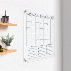 Minimalist acrylic wall calendar with black lettering, areas for notes, a menu, and a shopping list, with gold hardware.