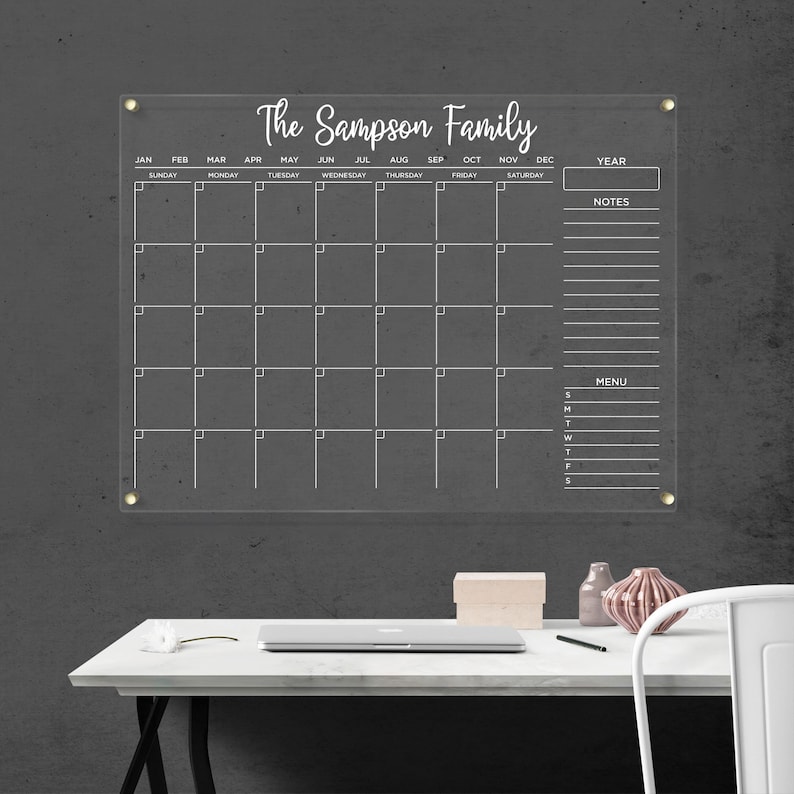 Clear acrylic wall calendar with gold hardware, featuring a full monthly layout with white writing, sections for year box, notes, and menu, displayed on a bright wall next to a decorative shelf.