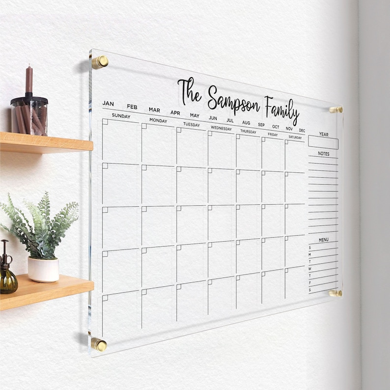 Clear acrylic wall calendar with gold hardware, featuring a full monthly layout, sections for year box, notes, and menu, displayed on a bright wall next to a decorative shelf.