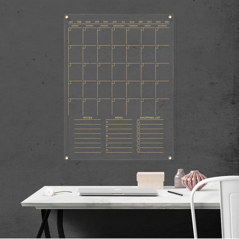 Minimalist acrylic wall calendar with gold lettering, areas for notes, a menu, and a shopping list, with gold hardware.