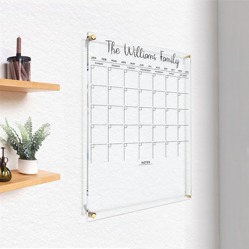 Clear acrylic wall calendar with gold hardware, showcasing a simple, elegant monthly layout with a notes section, displayed in a cozy corner with home decor.