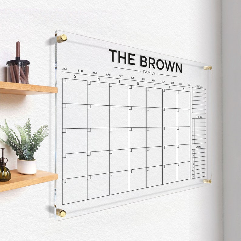 Acrylic family calendar with gold hardware and black lettering, including sections for monthly planning, notes, tasks, and menu.