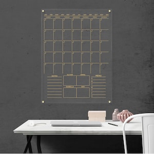 Featuring a monthly layout, notes, a menu section, and personalized spaces for four name boxes. Clear acrylic wall calendar with gold hardware and gold text.