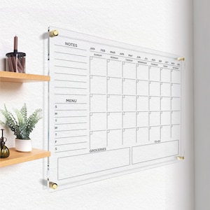 Clear wall calendar with black writing, featuring a notes column, menu section, grocery list, and to-do area, all outlined and mounted with gold hardware in a bright, organized space.