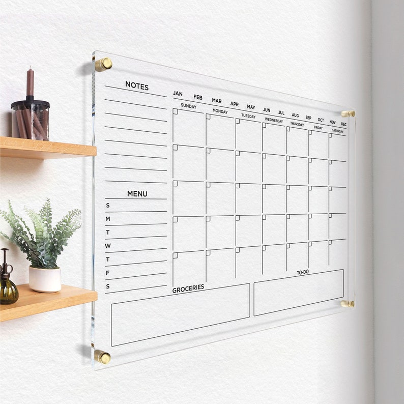 Clear wall calendar with black writing, featuring a notes column, menu section, grocery list, and to-do area, all outlined and mounted with gold hardware in a bright, organized space.