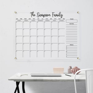 Clear acrylic wall calendar with gold hardware, featuring a full monthly layout with black writing, sections for year box, notes, and menu, displayed on a bright wall next to a decorative shelf.