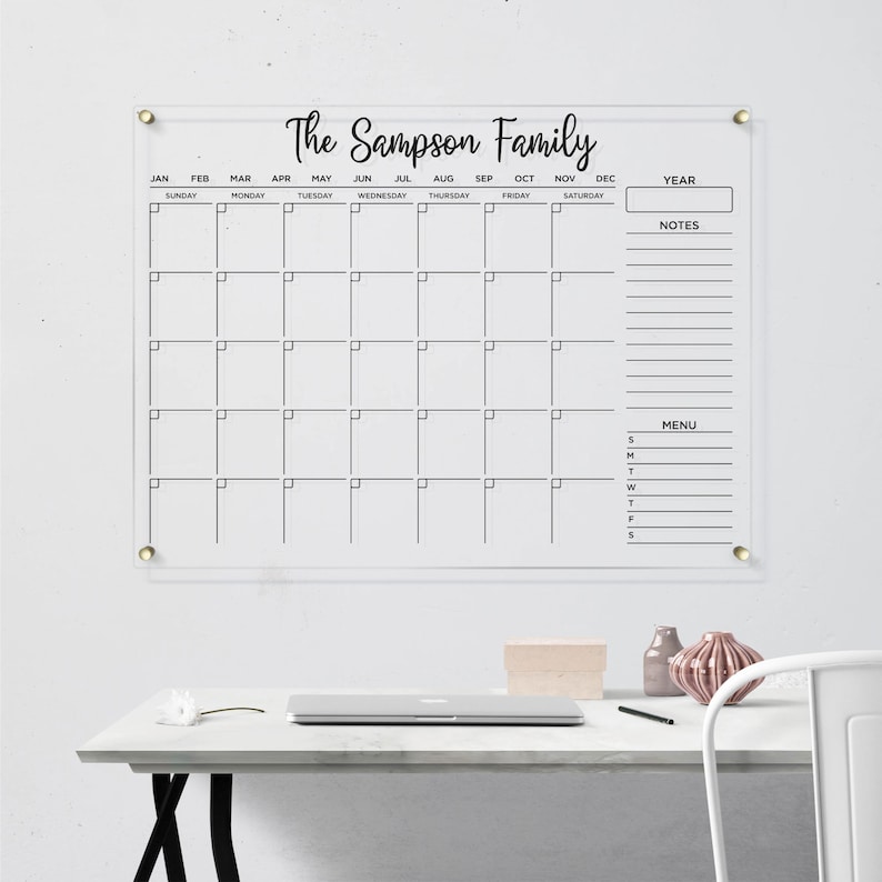 Clear acrylic wall calendar with gold hardware, featuring a full monthly layout with black writing, sections for year box, notes, and menu, displayed on a bright wall next to a decorative shelf.