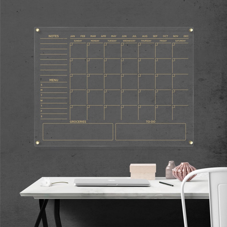 Clear wall calendar with gold writing, featuring a notes column, menu section, grocery list, and to-do area, all outlined and mounted with gold hardware.