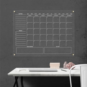 Clear wall calendar with white writing, featuring a notes column, menu section, grocery list, and to-do area, all outlined and mounted with gold hardware.