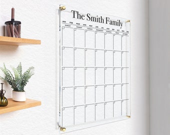 May include: A clear acrylic calendar with a black frame and gold screws. The calendar has the months of the year listed across the top and the days of the week listed down the side. The text "The Smith Family" is printed at the top of the calendar.