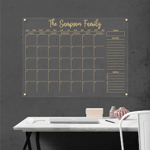 Clear acrylic wall calendar with gold hardware, featuring a full monthly layout with gold writing, sections for year box, notes, and menu, displayed on a bright wall next to a decorative shelf.