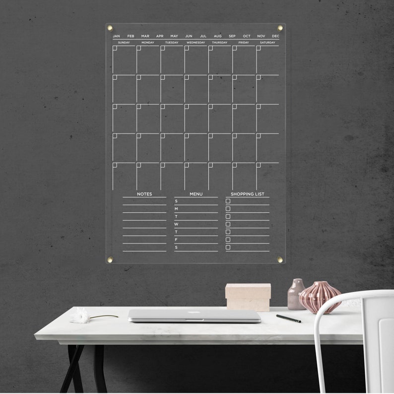 Minimalist acrylic wall calendar with white lettering, areas for notes, a menu, and a shopping list, with gold hardware.