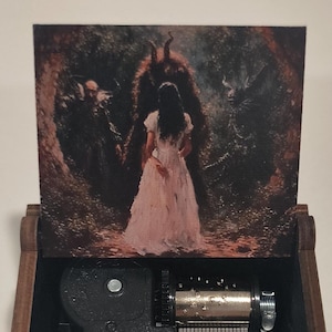 May include: A dark brown wooden music box with an open lid featuring a printed illustration of a figure in a white dress standing before two dark, horned entities. The interior contains a silver metal musical mechanism with a black plastic housing.