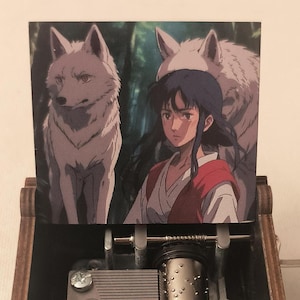 May include: A wooden music box with a hand-crank on the side. The lid is open and features a printed image of a woman with long black hair and two white wolves. The music box has a decorative design on the bottom.