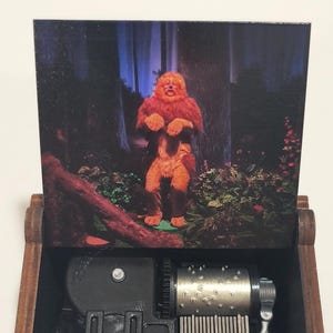 May include: A wooden music box with an open lid displaying a printed image of a furry orange creature standing in a dark forest. The interior features a silver metal cylinder with pins, a metal comb, and a black plastic winding mechanism.
