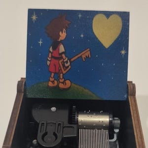 May include: A dark wooden music box with a hinged lid featuring a painted illustration of a character holding a key-shaped sword under a night sky with a yellow heart. The interior contains a mechanical metal music movement with a silver cylinder.