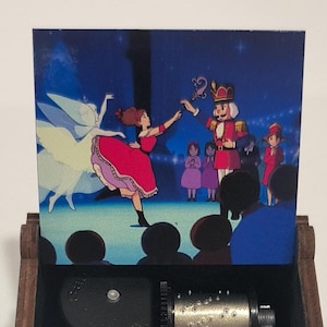 Puede incluir: Una caja de música de madera con tapa abatible, con una colorida ilustración de una escena de ballet de El Cascanueces. La caja está abierta, revelando los componentes mecánicos. El exterior es marrón oscuro con un diseño floral.