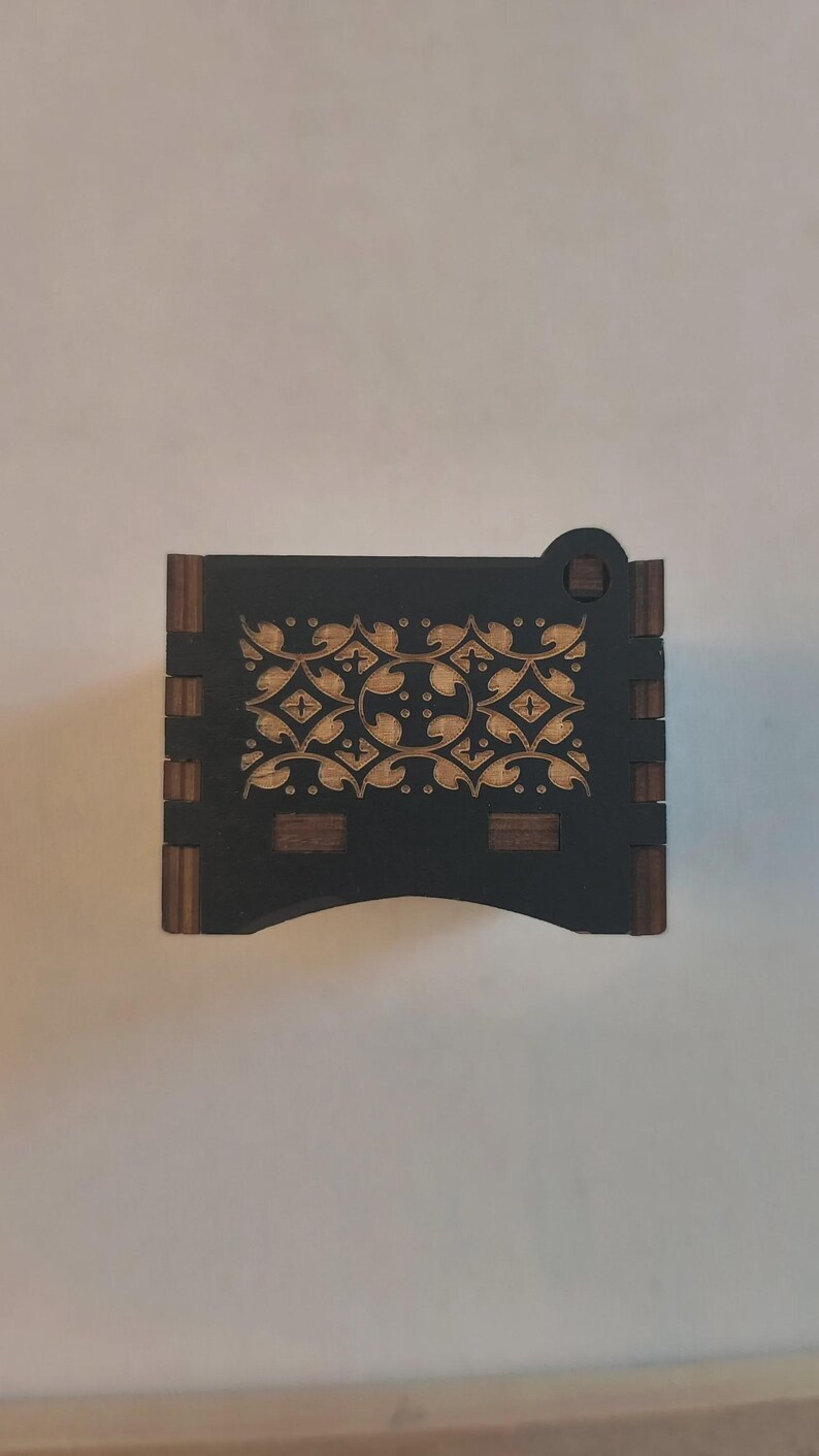 Pode incluir: Uma caixa retangular de madeira com acabamento preto e detalhes em madeira natural nas bordas. A superf&iacute;cie superior apresenta um padr&atilde;o floral intrincado gravado a laser em cor bege claro, contrastando com o fundo preto escuro.