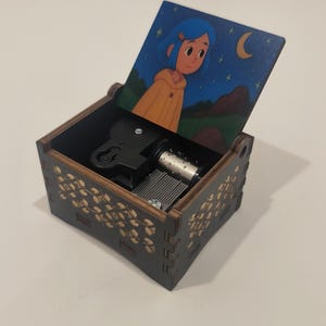 Pode incluir: Uma caixa de m&uacute;sica de madeira castanho-escura com tampa articulada com uma ilustra&ccedil;&atilde;o de uma pessoa de cabelo azul com um casaco amarelo sob um c&eacute;u noturno. Os lados exteriores t&ecirc;m um padr&atilde;o floral dourado e o mecanismo &eacute; vis&iacute;vel.