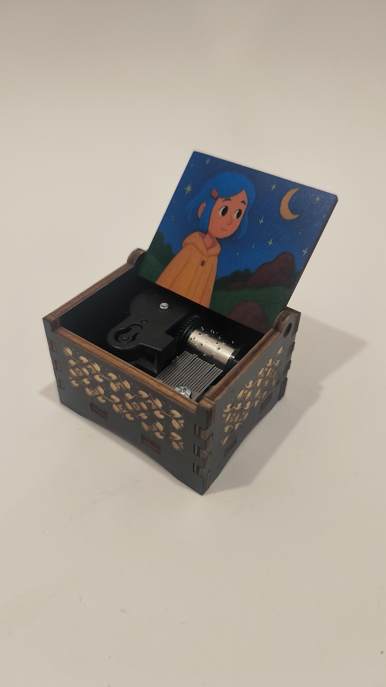 Pode incluir: Uma caixa de m&uacute;sica de madeira castanho-escura com tampa articulada com uma ilustra&ccedil;&atilde;o de uma pessoa de cabelo azul com um casaco amarelo sob um c&eacute;u noturno. Os lados exteriores t&ecirc;m um padr&atilde;o floral dourado e o mecanismo &eacute; vis&iacute;vel.