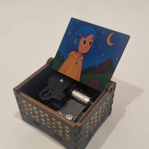 Pode incluir: Uma caixa de m&uacute;sica de madeira com acabamento castanho escuro e padr&otilde;es de folhas douradas no exterior. A tampa aberta apresenta uma ilustra&ccedil;&atilde;o colorida de uma personagem com cabelo azul usando um casaco amarelo sob um c&eacute;u noturno estrelado.