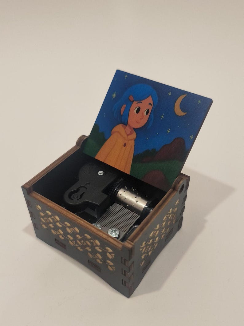 Pode incluir: Uma caixa de m&uacute;sica de madeira com acabamento castanho escuro e padr&otilde;es de folhas douradas no exterior. A tampa aberta apresenta uma ilustra&ccedil;&atilde;o colorida de uma personagem com cabelo azul usando um casaco amarelo sob um c&eacute;u noturno estrelado.
