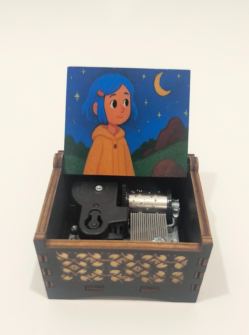 Pode incluir: Uma caixa de m&uacute;sica de madeira com exterior castanho escuro e um padr&atilde;o dourado de folhas. A tampa aberta exibe uma ilustra&ccedil;&atilde;o colorida de uma personagem de cabelo azul com um casaco amarelo sob um c&eacute;u noturno com lua e estrelas.