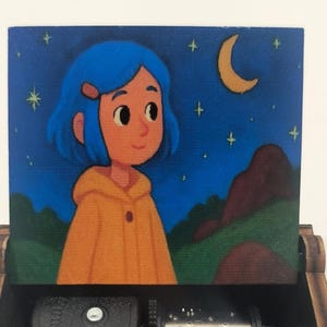 Pode incluir: Uma caixa de m&uacute;sica de madeira com uma tampa pintada apresentando uma personagem de cabelo azul vestindo um casaco amarelo com capuz. A cena retrata um c&eacute;u noturno com uma lua crescente e estrelas amarelas sobre colinas verdes e rochas.