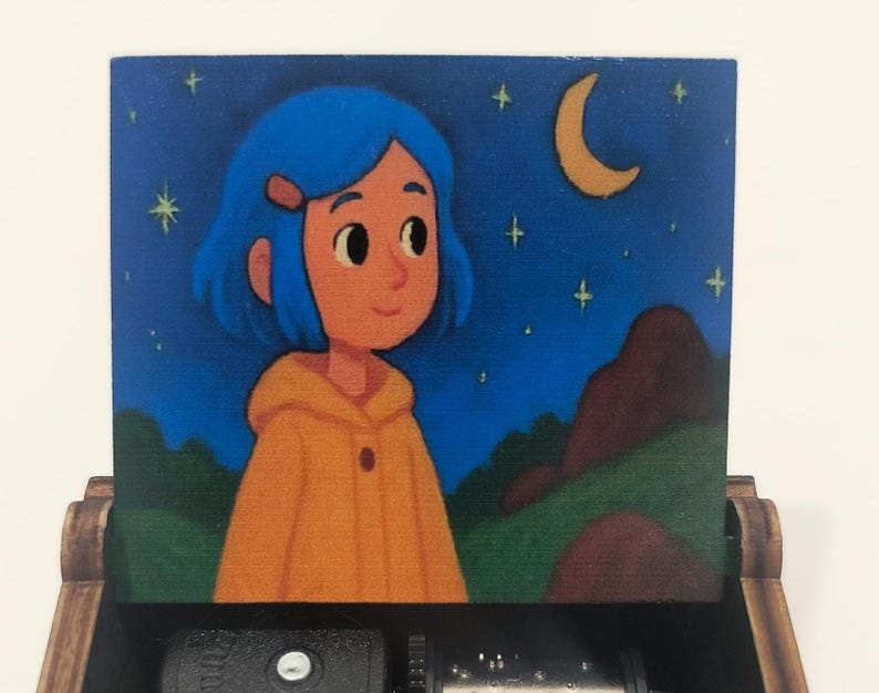 Pode incluir: Uma caixa de m&uacute;sica de madeira com uma tampa pintada apresentando uma personagem de cabelo azul vestindo um casaco amarelo com capuz. A cena retrata um c&eacute;u noturno com uma lua crescente e estrelas amarelas sobre colinas verdes e rochas.