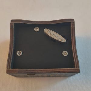 Pode incluir: Um mecanismo de caixa de m&uacute;sica quadrado de madeira com uma moldura castanha escura e uma base interior preta. O dispositivo inclui tr&ecirc;s parafusos de cabe&ccedil;a Phillips prateados e uma chave de corda de metal prateado com a marca Sankyo gravada.