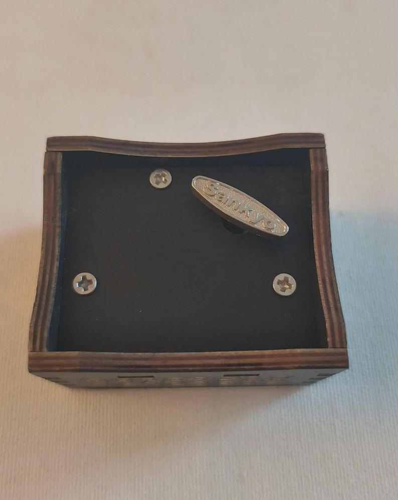 Pode incluir: Um mecanismo de caixa de m&uacute;sica quadrado de madeira com uma moldura castanha escura e uma base interior preta. O dispositivo inclui tr&ecirc;s parafusos de cabe&ccedil;a Phillips prateados e uma chave de corda de metal prateado com a marca Sankyo gravada.