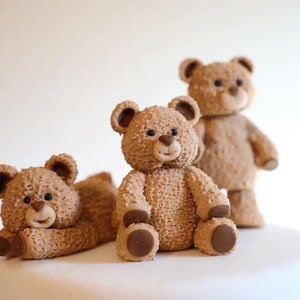 May include: Three brown teddy bear figurines on a white cake. The bears are made of fondant and have a fuzzy texture. The bears are in different poses, with one sitting up, one lying down, and one standing up.