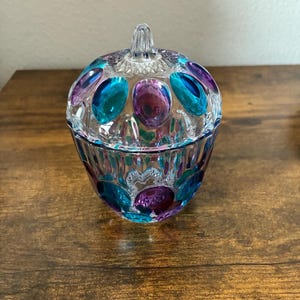 May include: A clear glass candy dish with a lid. The dish is decorated with teal, purple, and pink oval-shaped accents. The lid has a clear handle. The dish sits on a wooden surface.