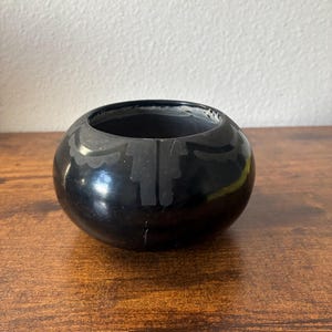 May include: A small, round, black ceramic bowl with a smooth, polished finish. The exterior features a matte black decorative band near the rim, consisting of repeating scalloped shapes and vertical rectangular patterns encircling the vessel.