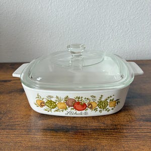 May include: A white rectangular casserole dish with a clear glass lid and integrated side handles. The exterior features a colorful botanical illustration of various vegetables and the text L'Echalote printed in a script font on the front side.