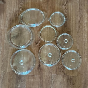 May include: A collection of clear glass cookware lids and dishes. The lids are round and oval, with small knobs. The dishes are oval and round. The items are arranged on a wooden surface.